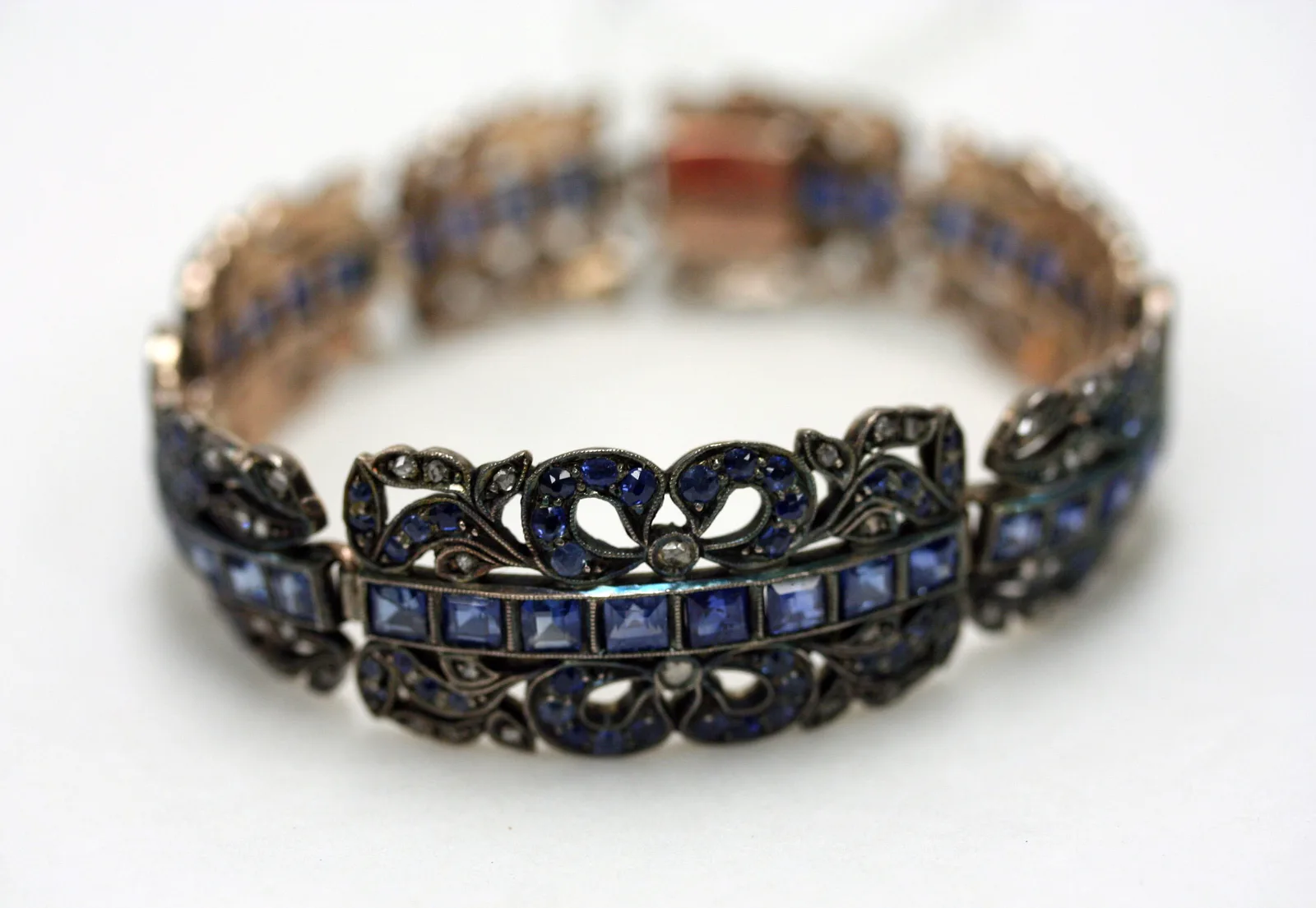 Austrian gold bracelet of rectangular links, each with a horizontal row of square sapphires set edge to edge in a metal trench, surrounded by diamond scrollwork