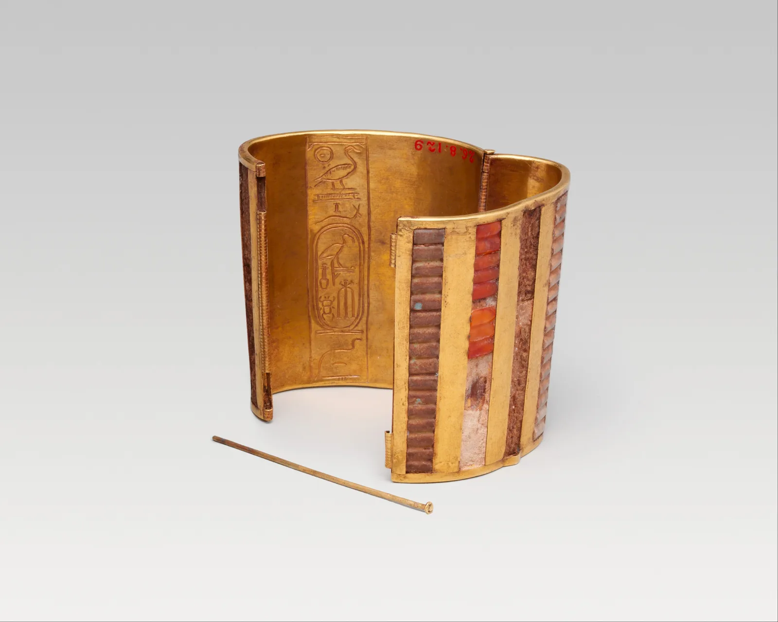 New Kingdom hinged gold cuff standing slightly open, its exterior ribbed into vertical trenches filled with rectangular carnelian, a gold pin beside the clasp