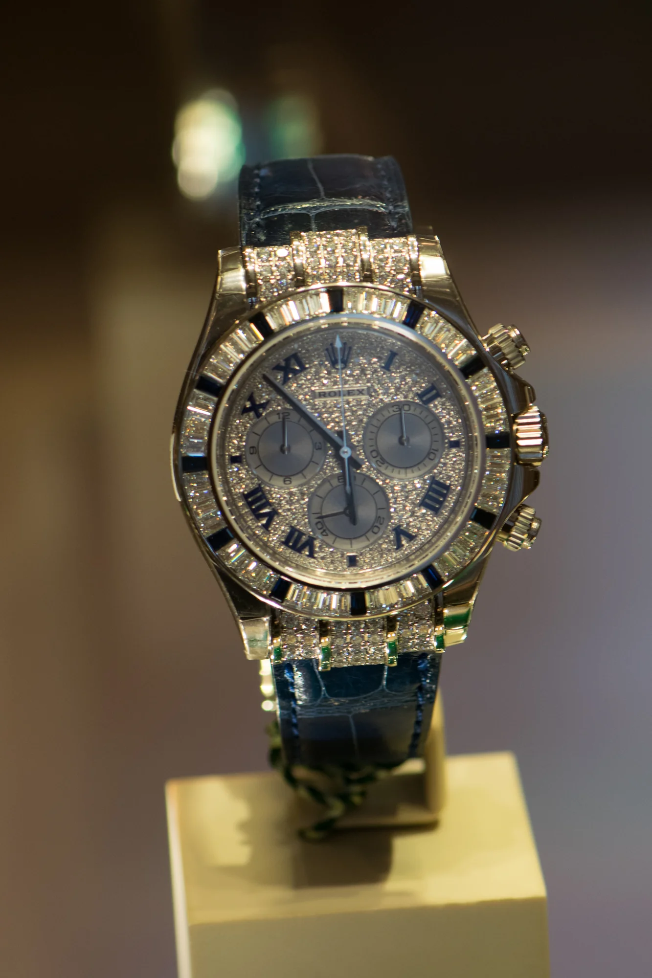 White-metal Rolex Daytona with a pavé diamond dial, baguette-diamond bezel with sapphire markers, and a dark leather strap, shown off the wrist on a stand