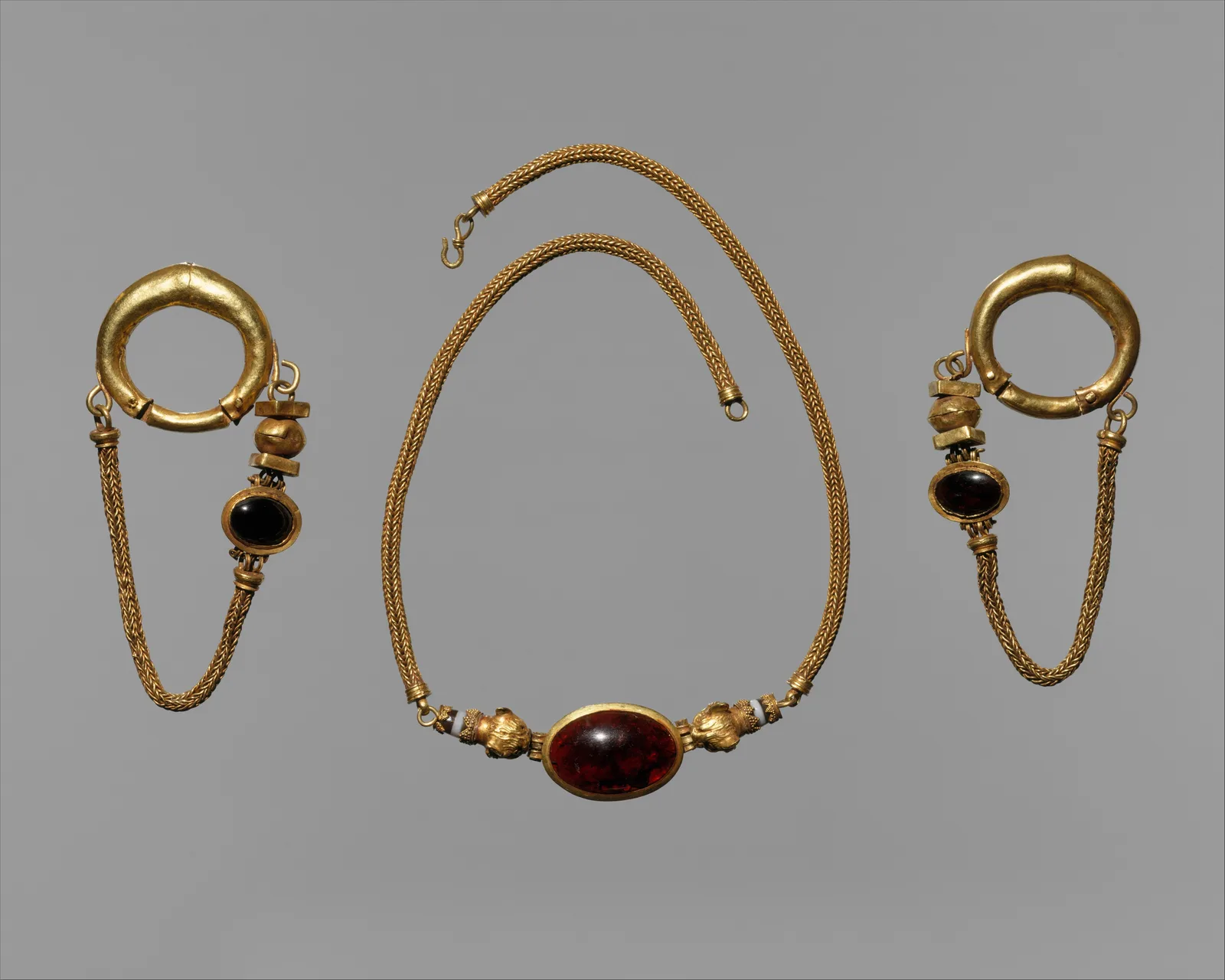 Late Hellenistic gold necklace with a large oval cabochon garnet at the center between lion-head terminals, shown with a pair of matching gold hoop earrings each hung with a smaller garnet