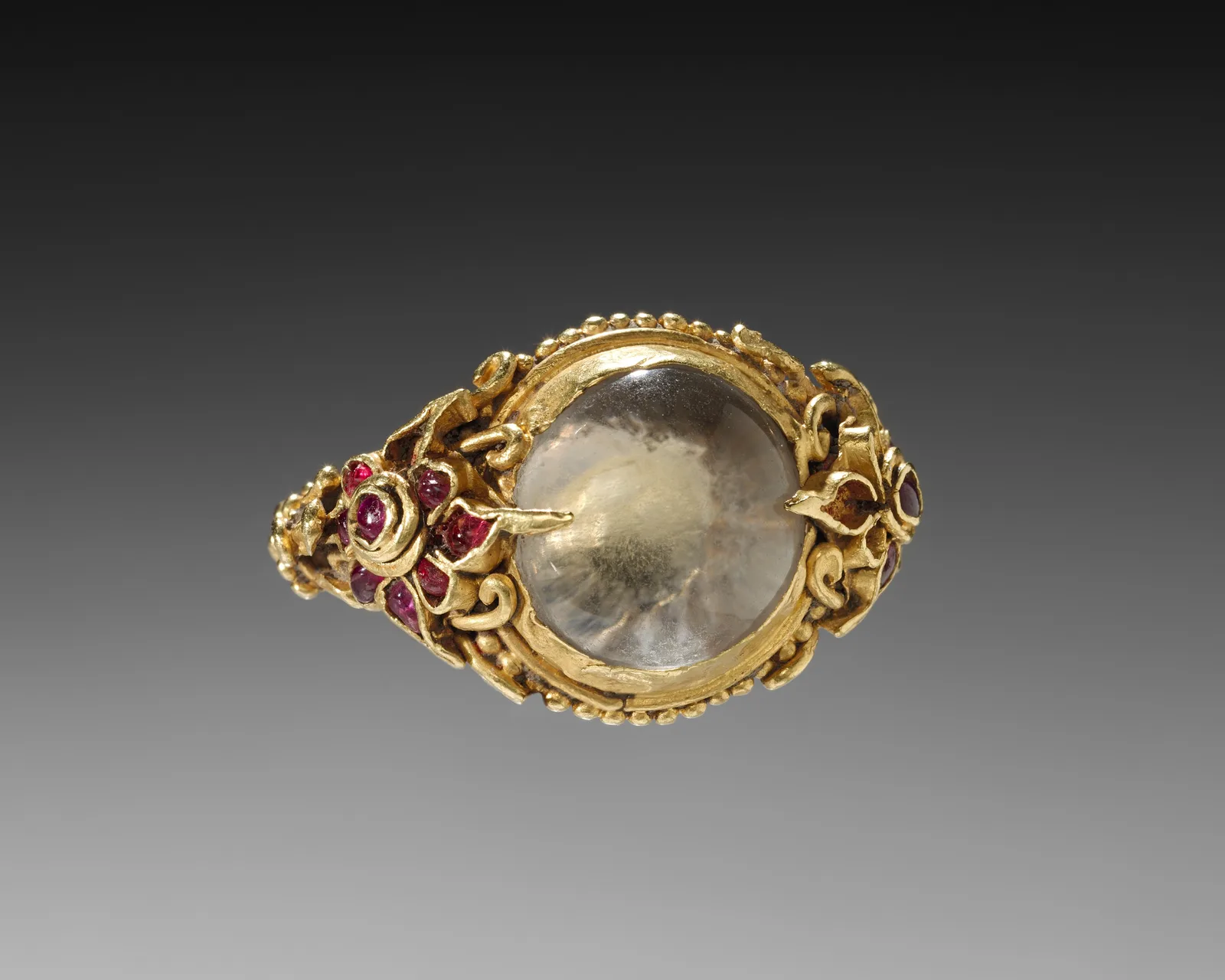Cambodian gold ring with a large domed moonstone in a granulated bezel, ruby clusters on the scrolling shoulders