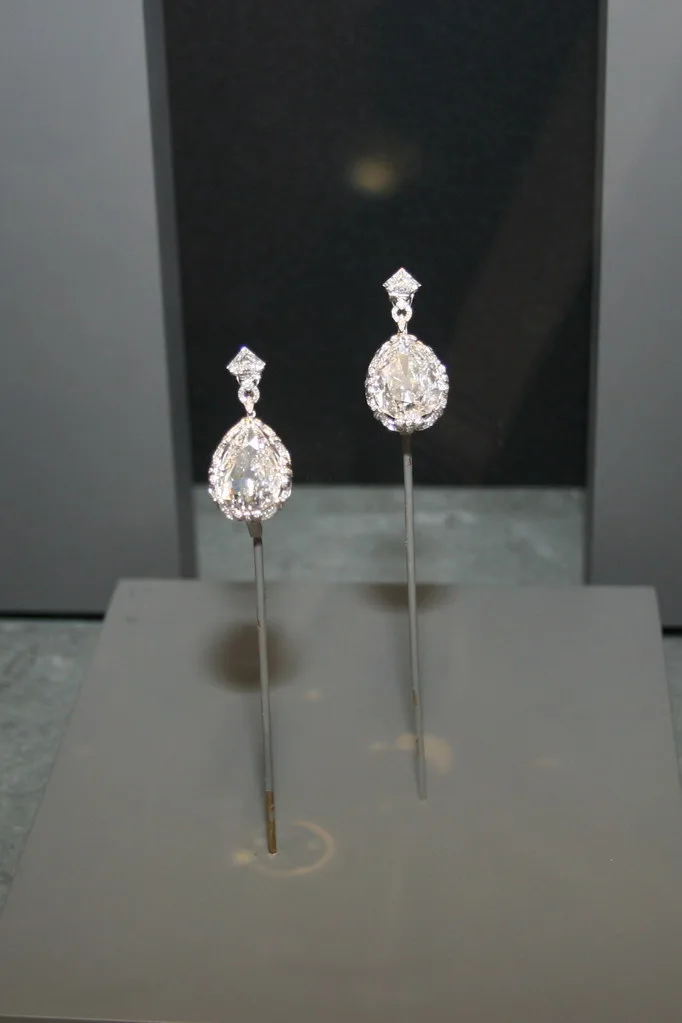 Pair of diamond drop earrings, each a pear-shaped stone in a dark museum display