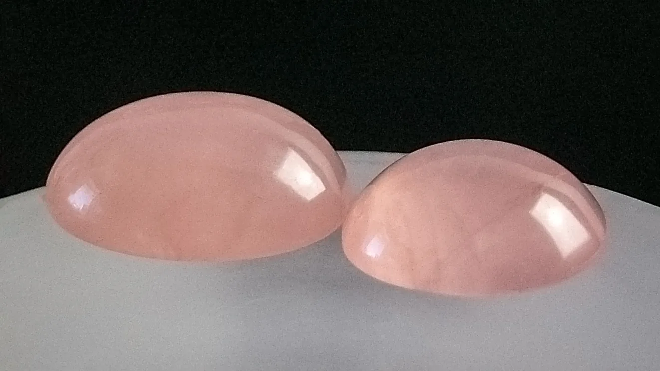 Two oval rose quartz cabochons, milky pink and softly translucent, on a clear glass against a pale ground