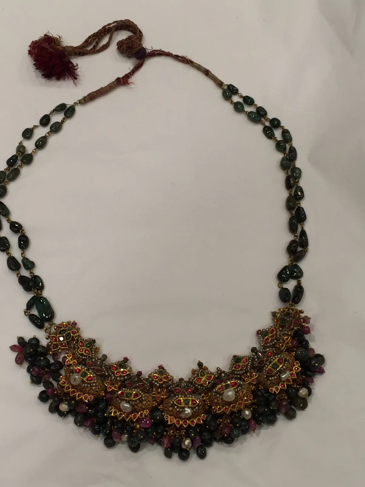 Northern Indian gold kaliganthi necklace laid as a crescent bib of filigree pendants set with red spinels and pearls, emerald bead strands, and a hanging fringe of green and red beads