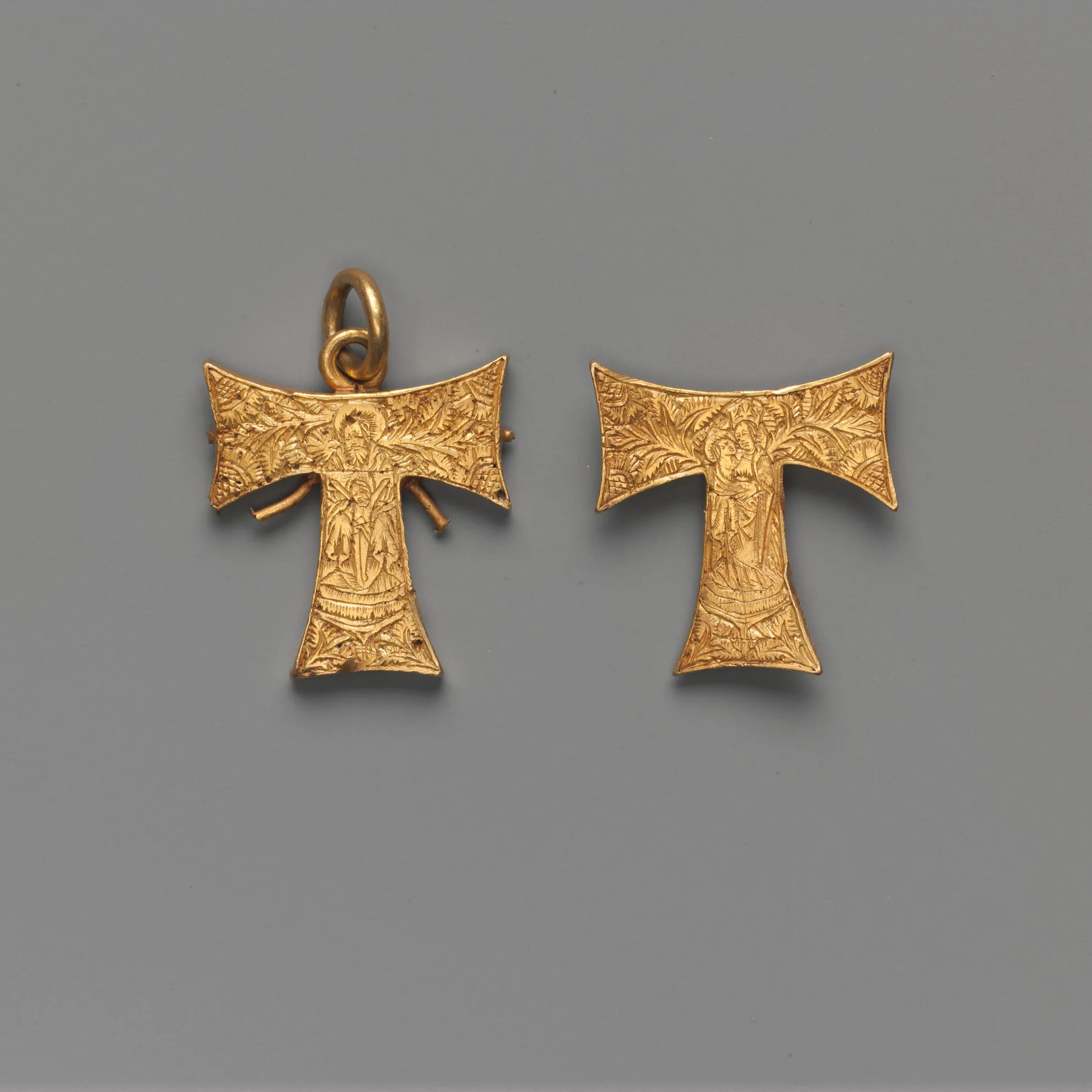 Two late-medieval gold pendants shaped as tau crosses, each a clear T of engraved gold with a standing figure on the stem