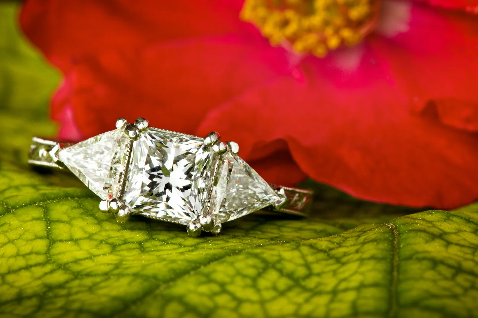 A gold ring with a princess-cut diamond at the center and a trilliant-cut diamond on each side, on a white ground