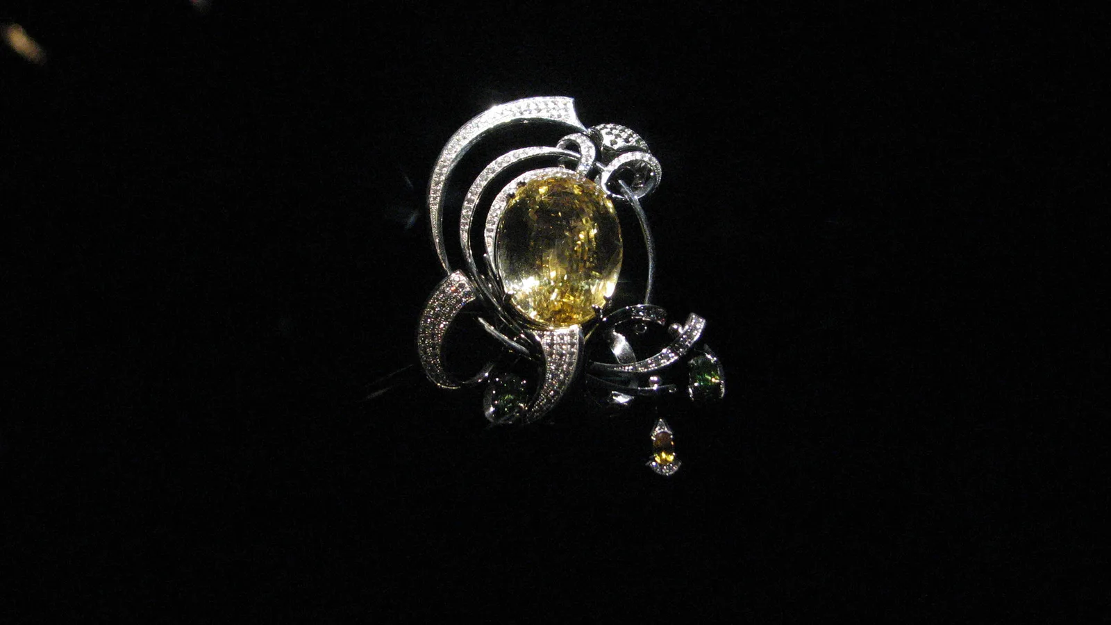 A platinum brooch centered on a large yellow sapphire, with white and green diamonds, on a black ground